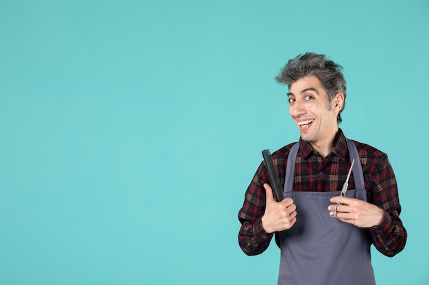 Close up view of smiling happy male barber wearing gray apron and holding comb scissor on blue color background