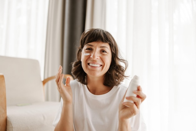 Крупный план улыбающейся женщины за кремом для кожи, смотрящей в камеру с бутылочным продуктом дома