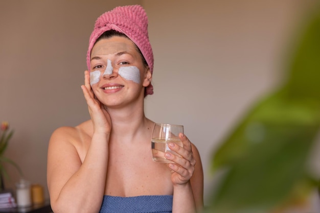 Close up view of smiling woman with cosmetic mask on face with glass water look at camera