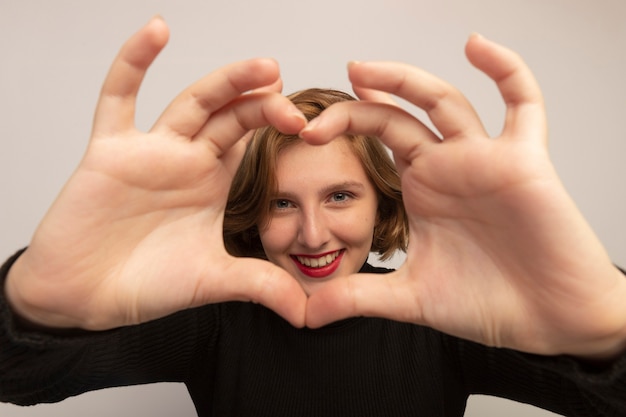Close-up view of smiling young blonde woman looking at front doing heart sign isolated on white wall