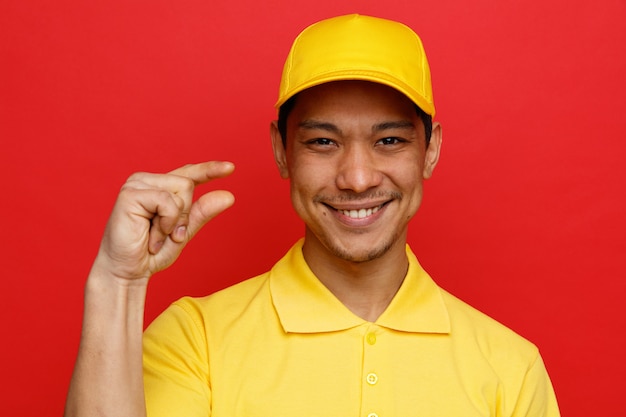 Close-up view smiling young delivery man wearing uniform and cap doing small amount gesture 