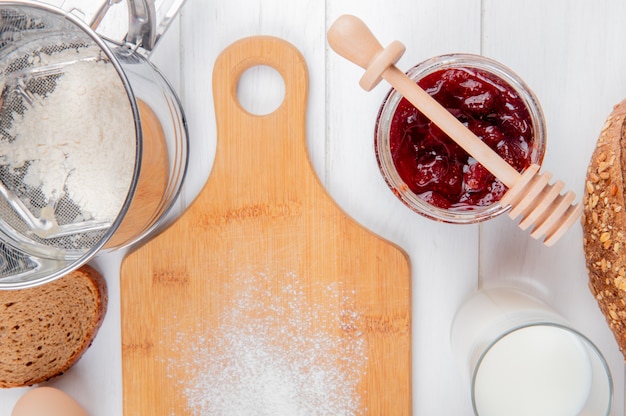 Close-up view of strawberry jam in jar flour slice of rye bread glass of milk and cutting board on wooden surface