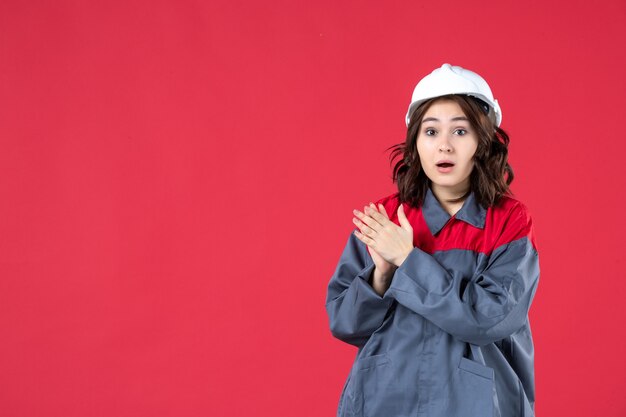 Close up view of surprised female builder in uniform with hard hat and applausing someone on isolated red wall