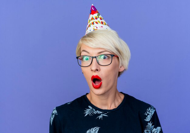 Free photo close-up view of surprised young blonde party girl wearing glasses and birthday cap looking at camera isolated on purple background with copy space