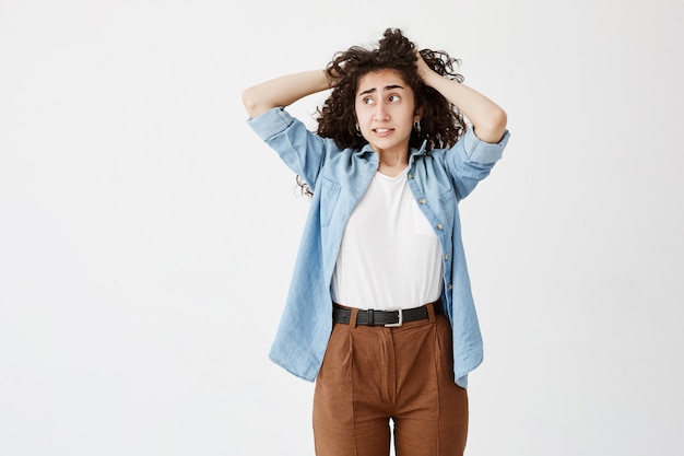 Close up view of teenage girl in denim shirt and brown trousers, looking aside with puzzled face expression, clenches teeth, touching her long dark wavy hair. Face expression and emotions concept