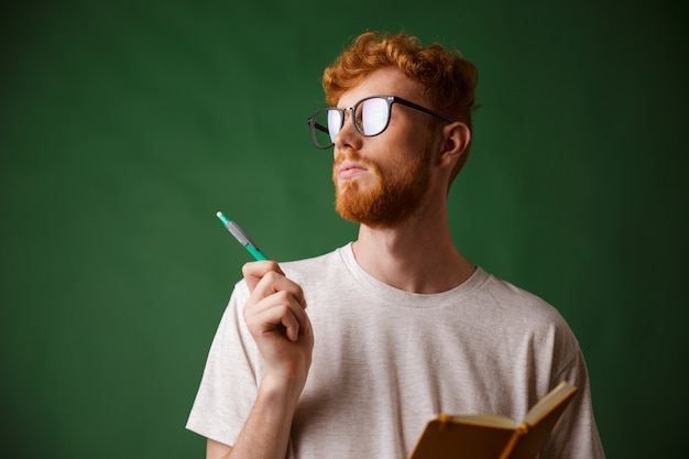 Close-up view of thinking bearded young man in white tshirt holding a notebook and a pen