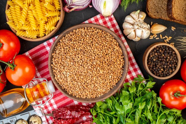 Close up view of uncooked buckwheat in a brown bowl on towel fresh vegetables fallen oil bottle salt green bundle dried pepper