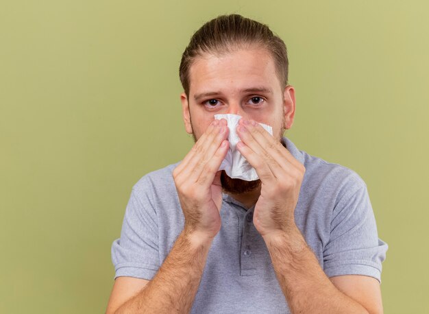 オリーブグリーンの壁に隔離された正面を見てナプキンで彼の鼻を拭く弱い若いハンサムな病気の男の拡大図