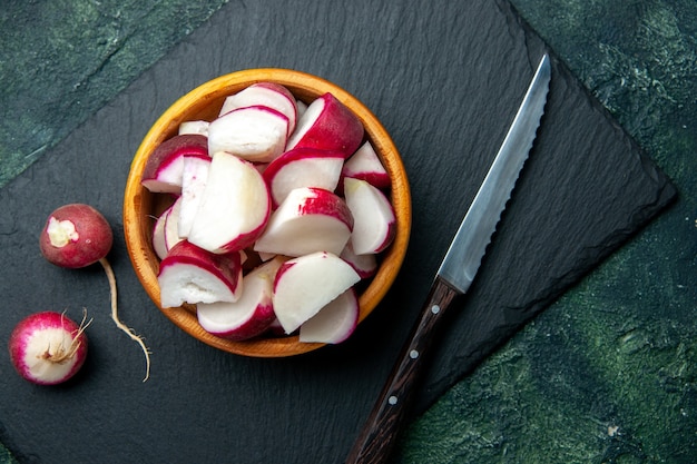 Close up view of whole and chopped fresh radishes in bowl and knife on dark color cutting board on green black mixed colors background with free space