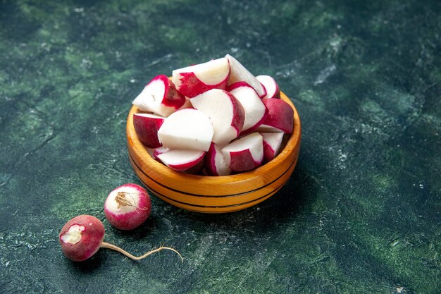 Close up view of whole and chopped fresh radishes inside and outside of a wooden bowl on dark color background with free space