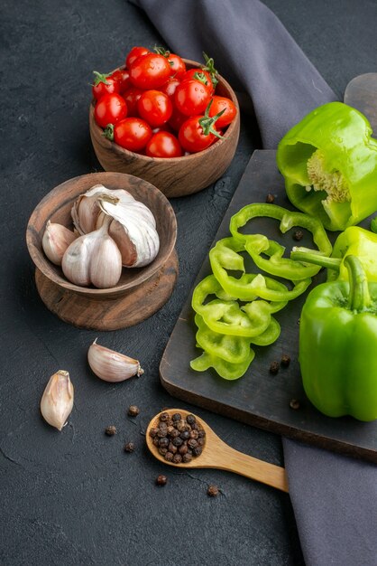 Close up view of whole cut chopped green peppers on wooden cutting board tomatoes in bowl garlics on dark color towel on black surface