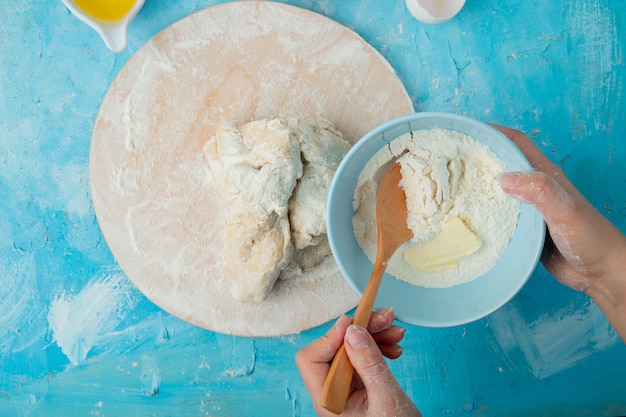 Close-up view of woman hand adding flour to dough on wooden surface and blue background
