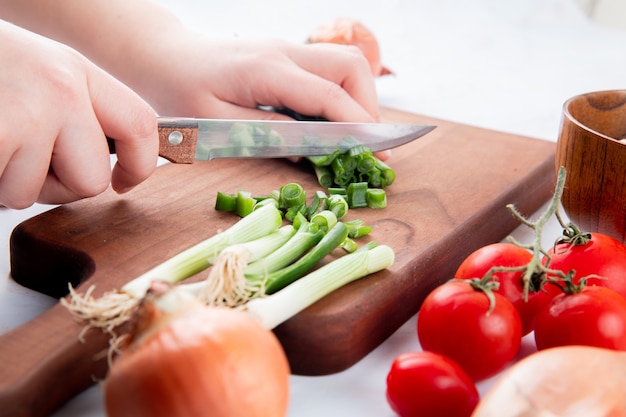 Close-up view of woman hand cutting green onion on cutting board with knife and tomatoes on white background