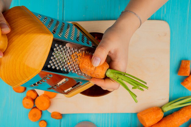 Close-up view of woman hands grating carrot on metal grater with cutting board and carrots on blue background