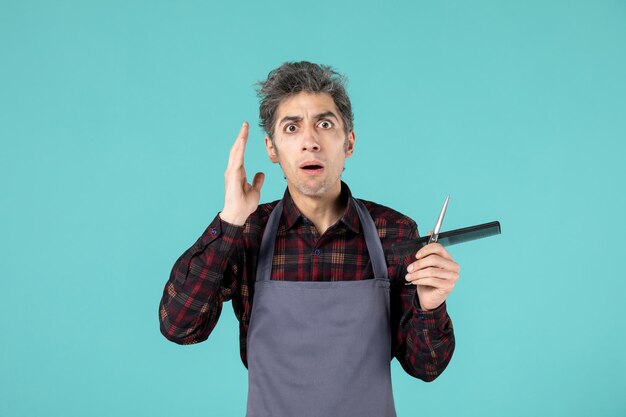 Close up view of young confused emotional male hairdesser wearing gray apron and holding scissor comb on pastel blue color background