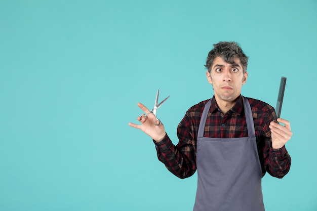 Close up view of young confused male barber wearing gray apron and holding scissors and comb on soft blue color background