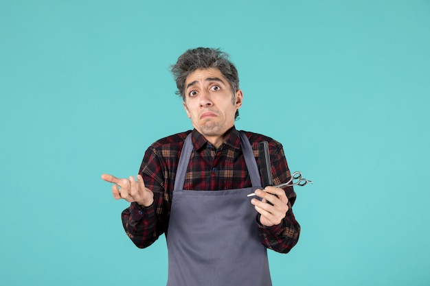 Close up view of young confused male hairdesser wearing gray apron and holding scissor comb on pastel blue color surface