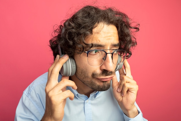 Vista ravvicinata di giovane uomo caucasico bello con gli occhiali e le cuffie guardando lato ascoltando musica isolata sulla parete cremisi