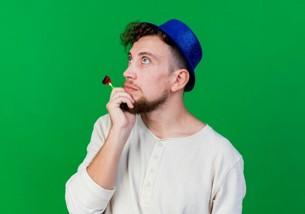 Close-up view of young handsome slavic party guy wearing party hat holding party blower touching chin looking up isolated on green wall