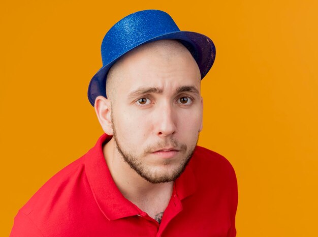 Close-up view of young handsome slavic party guy wearing party hat looking at front isolated on orange wall with copy space