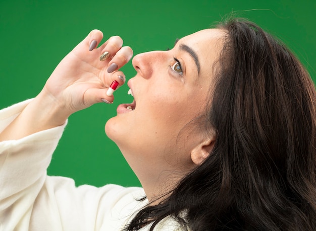 Free photo close-up view of young ill woman wearing robe standing in profile view taking pill looking at side isolated on green wall