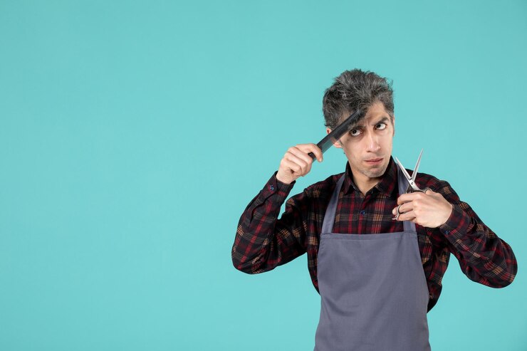 मुफ़्त फोटो close up view of young serious male barber wearing gray apron and combing his hair on pastel blue color surface