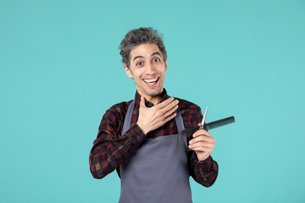 Close up view of young smiling happy male hairdesser wearing gray apron and holding scissor comb on pastel blue color background