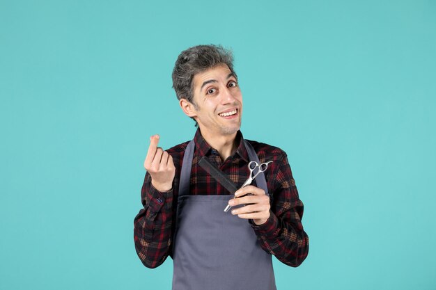 Close up view of young smiling male hairdesser wearing gray apron and holding scissor comb on pastel blue color background