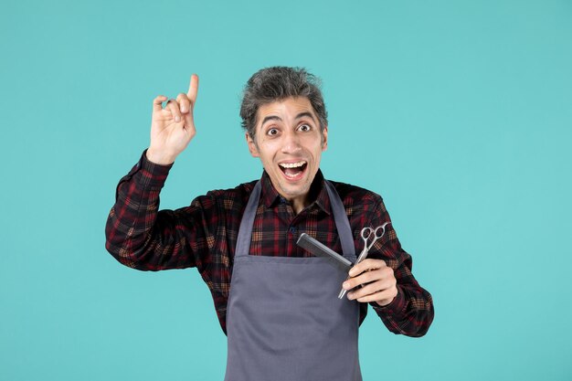 Close up view of young surprised male hairdesser wearing gray apron and holding scissor comb pointing up on pastel blue color background