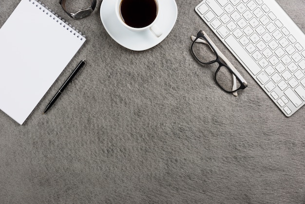 Close-up of white coffee cup; keyboard; wrist watch; pen; spiral notepad; eyeglasses and keyboard on desk