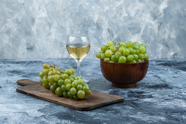 ボウルに白ブドウとグラスワイン、濃い水色の大理石の背景にまな板の上にブドウをクローズアップ。水平