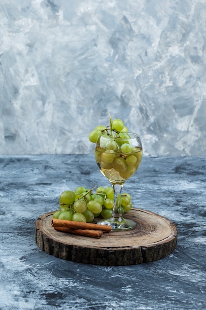 Close-up white grapes, cinnamon, glass of whisky on wooden board on dark blue marble background. vertical