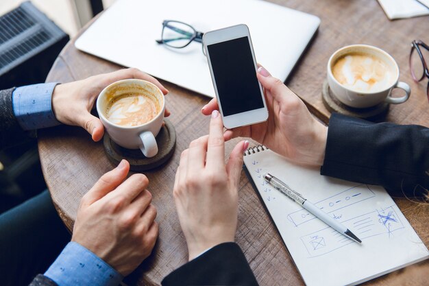 Close-up of white smartphone in woman's hands. Two colleagues sitting at a coffee table drinking coffee