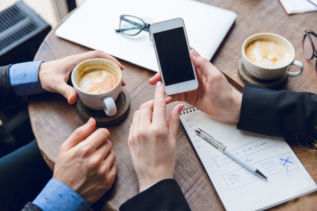 Крупный план белого смартфона в руках женщины. Двое коллег сидят за журнальным столиком и пьют кофе