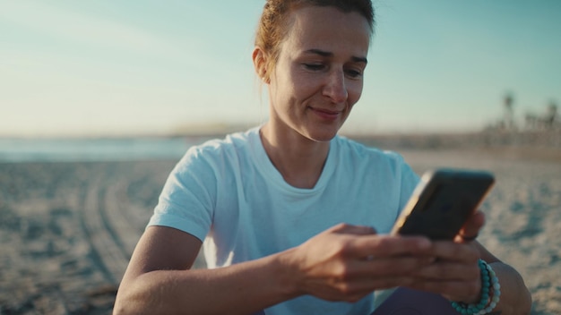 海のそばでスマートフォンを使用してソーシャルメディアをチェックしている女性をクローズアップビーチでのトレーニング後に休んでいるスポーティな女の子