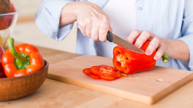 木製のまな板にナイフで新鮮な赤ピーマンを切る女性のクローズアップ