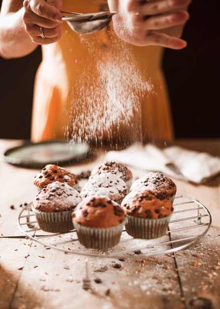 Close-up of a woman dusting icing sugar on muffins over the cooling rack
