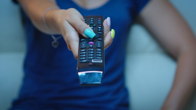 Close up of woman hand changing TV channels sitting on couch. Television remote control in the hands of lady pointing the TV and choosing a movie, holding controller and pressing the button