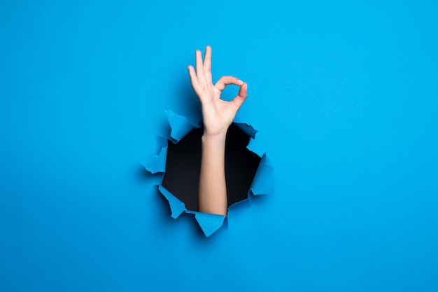 Закройте вверх руки женщины с одобренным жестом через голубое отверстие в бумажной стене.