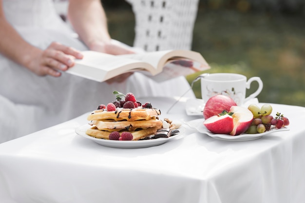 Крупным планом женщина, держащая книгу в руке, сидя за столом для завтрака на открытом воздухе
