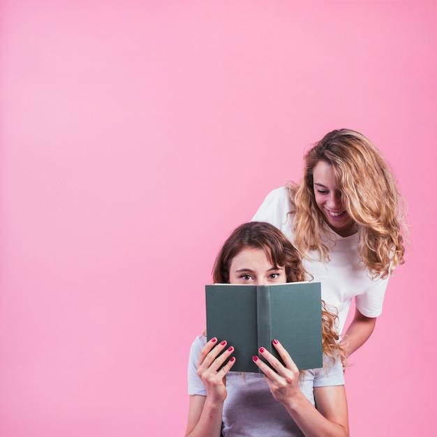 Close-up of woman looking at her friend reading book against pink background