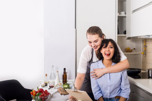 Close-up of a woman loving his friend looking at recipe on clipboard in the kitchen