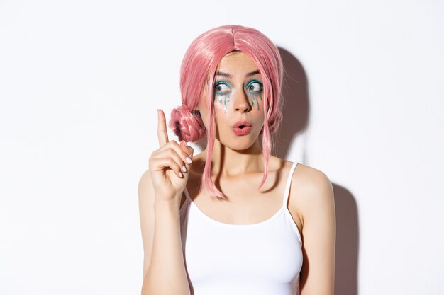 Close-up of woman in pink party wig and bright makeup, have idea, raising index finger in eureka sign, standing.
