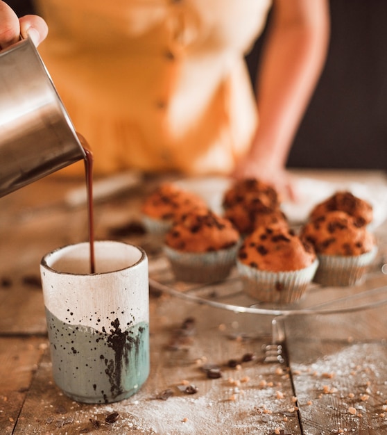 Close-up of a woman pouring melted chocolate in glass with fresh muffin on cooling rack