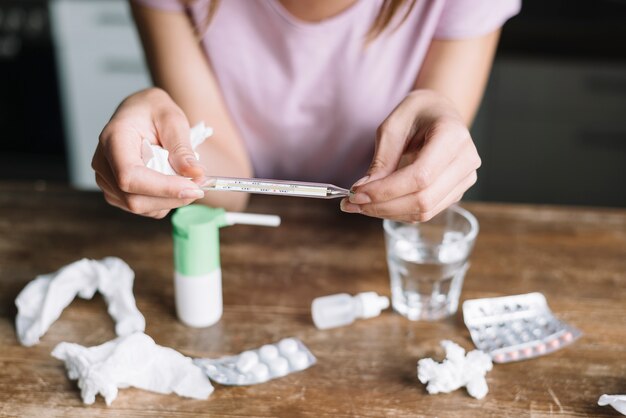 Close-up of a woman's hand checking temperature on thermometer with medicines on wooden desk