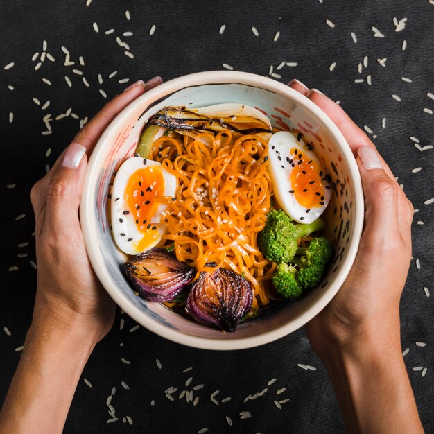 Close-up of woman's hand holding bowl of noodles with eggs; onion; broccoli in bowl on black background