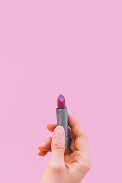 Close-up of a woman's hand holding dark shade lipstick over pink backdrop