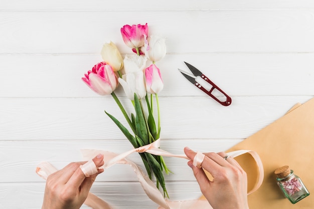 白い机の上のリボンとカッターを使用しながらチューリップの花の花束を作る女性の手のクローズアップ