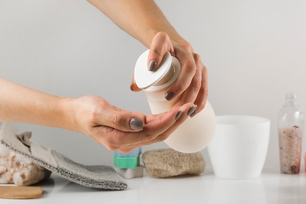 Close-up of a woman's hand pouring sanitizer soap from dispenser with spa product on white desk against background