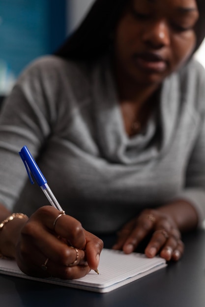 Close up of woman taking notes with pen on textbook at desk. Young adult writing information on notebook papers while working from home on business project. Person with remote work